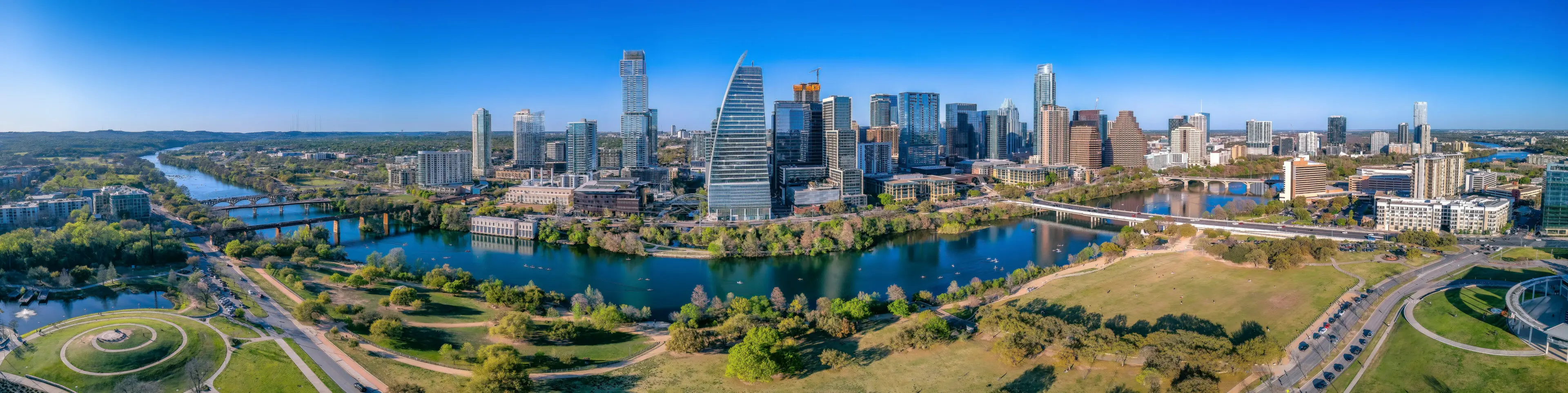 Panoramic view of Colorado River and Austin Texas cityscape. There is a view of a park at the front and bridges over the river near the skyscrapers.